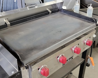 May include: A stainless steel flat top griddle with a black base and red knobs. The griddle has a large flat surface and is on a stand with wheels.