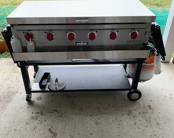 May include: A stainless steel flat top grill with six red knobs and a black metal cart. The grill has the words "Flat Top Grill" and "Samac Craft" printed on the front.