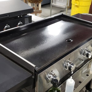 May include: A stainless steel flat top grill with a black griddle surface and multiple control knobs. The grill has a side shelf and a built-in handle.