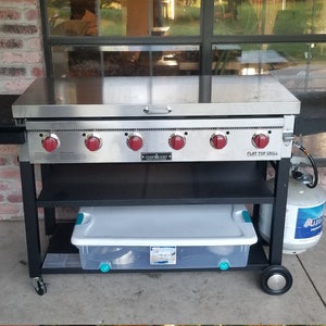 May include: A black and stainless steel flat top grill with five red knobs and a side shelf. The grill is on a black cart with a shelf and wheels. A propane tank is next to the cart.