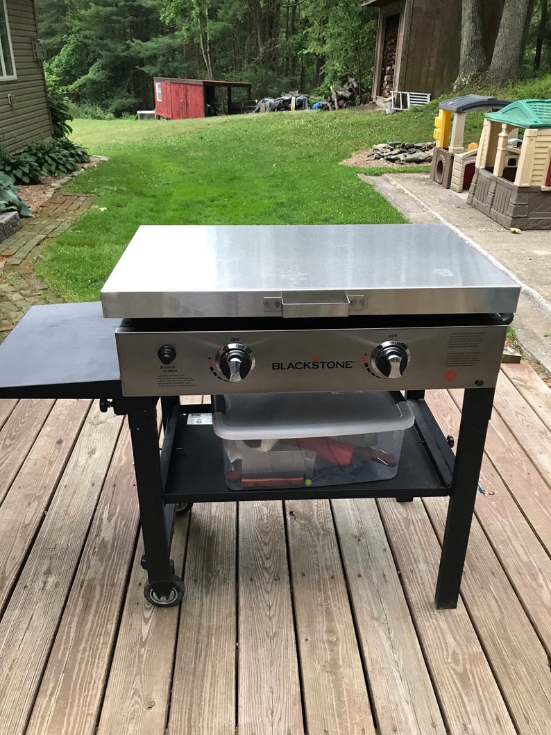 May include: Blackstone flat top grill with two burners and a side table. The grill is on a black metal stand with wheels. The grill is on a wooden deck.