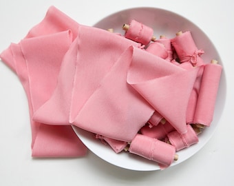 Puede incluir: Cintas de tela rosas, algunas enrolladas y otras planas, dispuestas en un cuenco blanco y sobre una superficie blanca. Las cintas son de un rosa pastel suave, y las enrolladas están sobre pequeños carretes de madera.