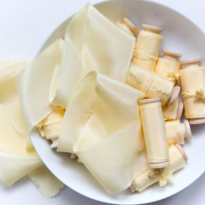 May include: Pale yellow silk ribbons and spools are displayed in a white bowl. The ribbons are loosely arranged, and the spools are wound with matching silk. The background is a bright white, creating a clean aesthetic.