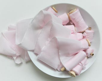 Puede incluir: Un cuenco blanco lleno de cinta de seda rosa pálido y carretes. La cinta está suelta y los carretes están cuidadosamente dispuestos. La cinta y los carretes son de un color rosa suave y delicado, creando una estética romántica y elegante.