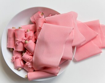 Puede incluir: Un tazón blanco lleno de carretes de cinta de seda rosa. La cinta está dispuesta de forma suelta y fluida, con parte de la cinta que se derrama del tazón.