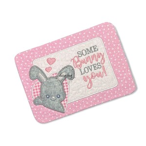 May include: A pink fabric square with a white border and a pink and white heart design. The fabric square has a white rectangle with the text "Some Bunny Loves You!" and a grey bunny with pink ears and a pink heart.