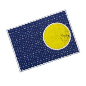 Pode incluir: Um tapete de mesa azul de tecido com uma borda branca costurada e um círculo amarelo no centro. O círculo tem o texto "daisy bay" em branco.