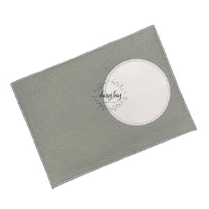 以下が含まれることがあります： 中央に白い円がある灰色のフェルトのクラフトプロジェクト。円は白いステッチの縁取りがされています。円の中央には「daisy kay embroidery」というテキストがあります。