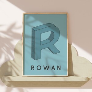 May include: A framed print with a blue background featuring a 3D stylized letter "R" in a darker blue color. The name "ROWAN" is printed in black below the letter.