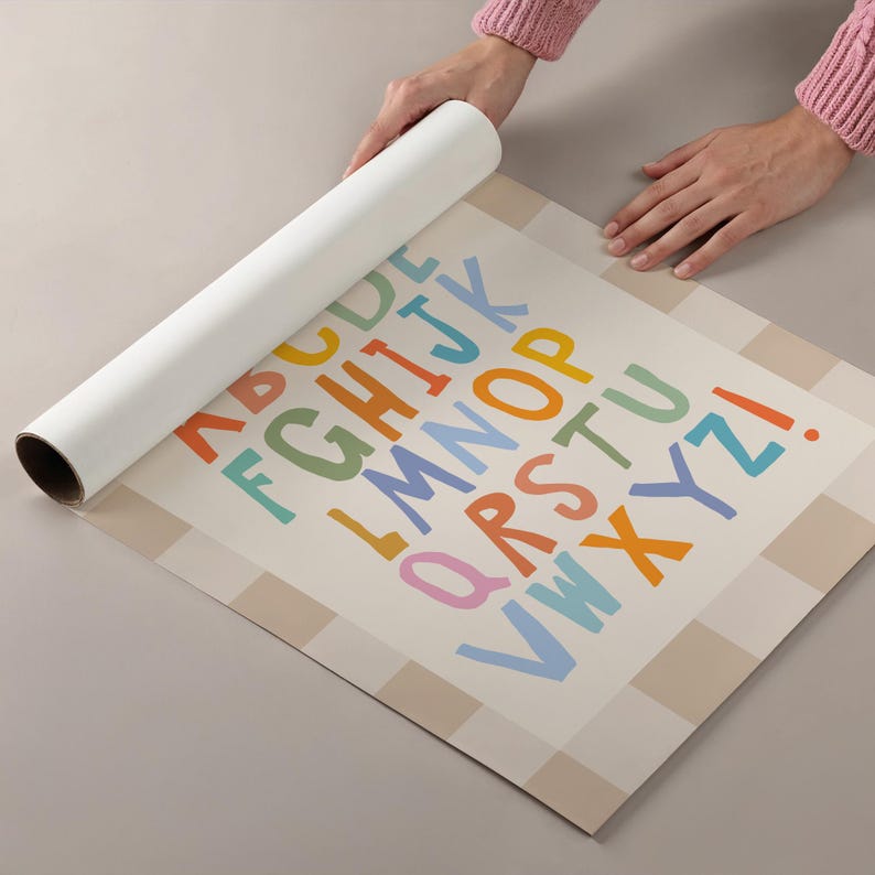 Puede incluir: Un p&oacute;ster enrollado que presenta el alfabeto en una fuente colorida de estilo manuscrito. Las letras de la A a la Z y un signo de exclamaci&oacute;n est&aacute;n impresas en tonos naranja, azul, verde, amarillo y rosa sobre un fondo beige a cuadros.