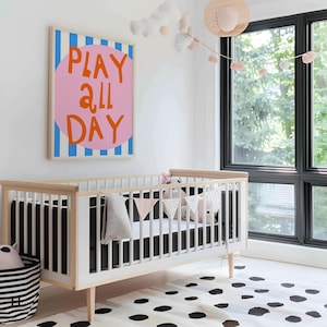 May include: A nursery scene featuring a white crib with a black mattress and a decorative banner. A large framed artwork with the words "PLAY ALL DAY" in orange letters on a pink circle is mounted on the wall. A black and white striped basket is on the floor.