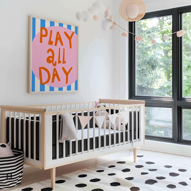 May include: A nursery scene featuring a white crib with a black mattress and a decorative banner. A large framed artwork with the words "PLAY ALL DAY" in orange letters on a pink circle is mounted on the wall. A black and white striped basket is on the floor.