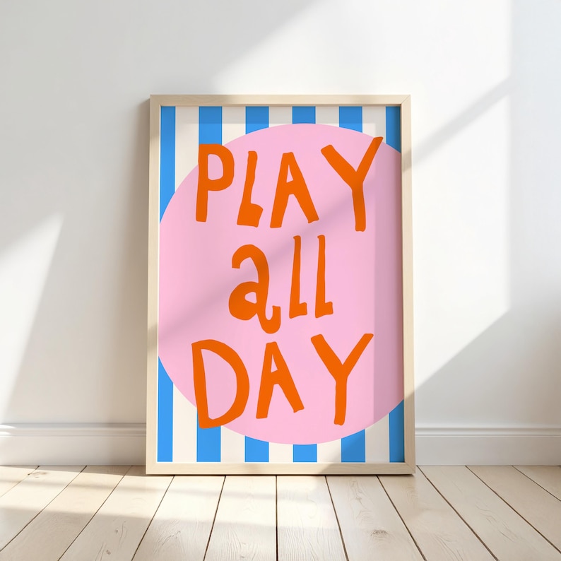 May include: A framed art print with the words "PLAY ALL DAY" in orange letters on a pink circle. The background has blue and white vertical stripes. The frame is a light wood color.