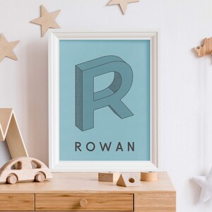 May include: A blue and white framed print with a large, 3D, blue letter "R" and the name "ROWAN" printed below it. The print is on a light blue background.
