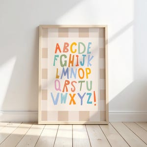 Puede incluir: Una l&aacute;mina de arte mural enmarcada que muestra el alfabeto con letras coloridas de estilo manuscrito dispuestas en filas. El fondo de la l&aacute;mina tiene un patr&oacute;n de cuadros en beige claro y blanco, con letras en tonos naranja, azul, verde y rosa.