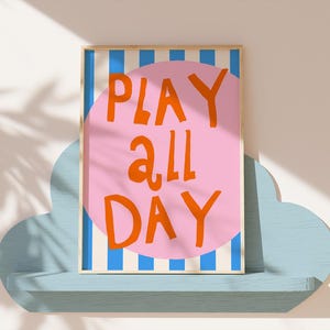 May include: A framed art print with the words "PLAY ALL DAY" in orange on a pink circle, set against a background of blue and white stripes. The artwork is displayed on a light blue cloud-shaped shelf.