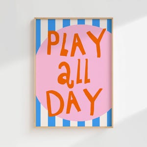May include: A framed art print with the words "PLAY ALL DAY" in orange, set against a pink circle. The background features blue and white vertical stripes. The frame is a light brown color.