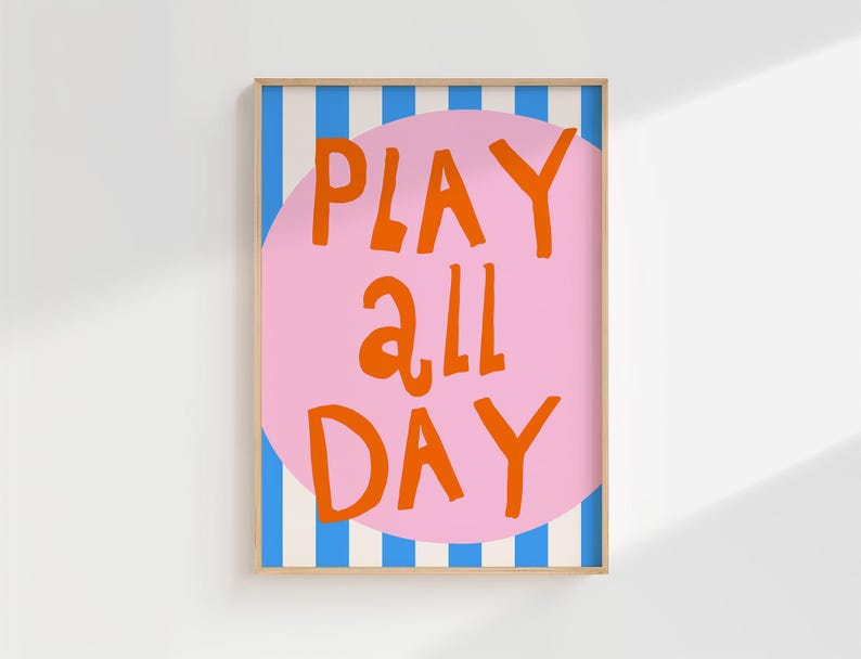 May include: A framed art print with the words "PLAY ALL DAY" in orange, set against a pink circle. The background features blue and white vertical stripes. The frame is a light brown color.