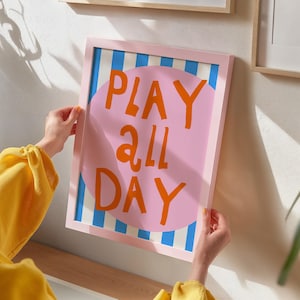 May include: A framed art print with the words "PLAY ALL DAY" in orange letters on a pink circle. The background has blue and white stripes. The frame is light pink.