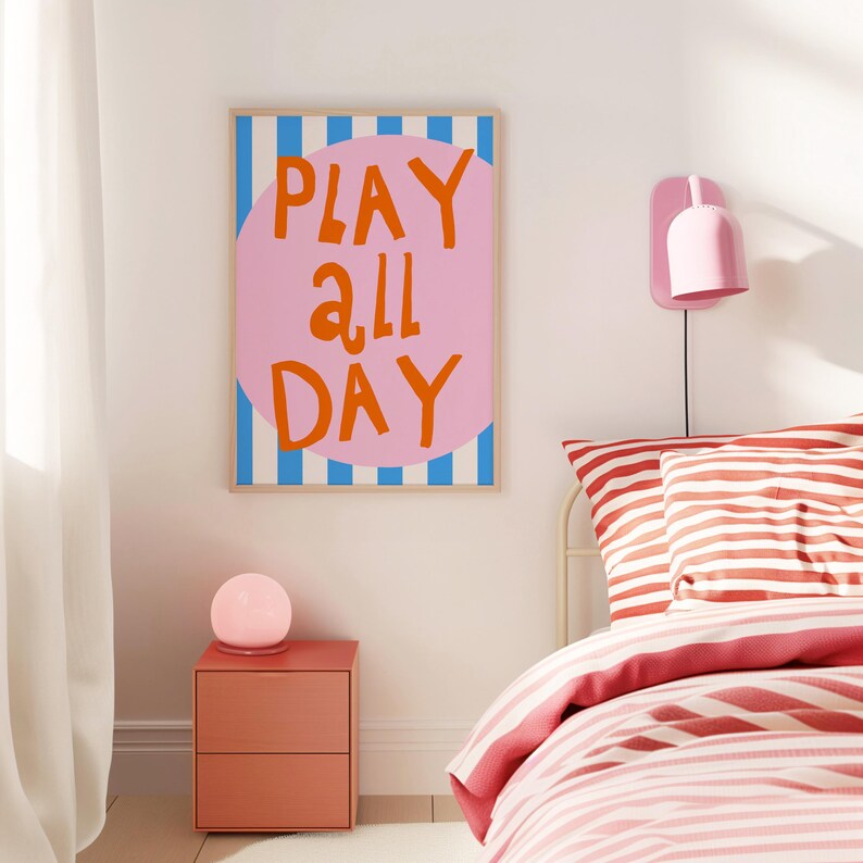 May include: A framed poster with the words "PLAY ALL DAY" in orange lettering on a pink circle, set against a blue and white striped background. The poster is in a bedroom with a matching pink nightstand and a bed with red and white striped bedding.
