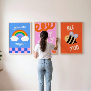 May include: Three framed wall art prints. The left print is blue with a rainbow, clouds, and a pink and blue checkerboard pattern with the text you are magic. The center print is pink with orange swirls. The right print is orange with a bee and the text BEE YOU.