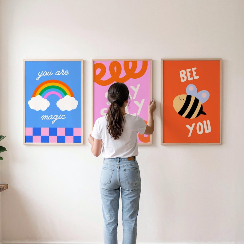 May include: Three framed wall art prints. The left print is blue with a rainbow, clouds, and a pink and blue checkerboard pattern with the text you are magic. The center print is pink with orange swirls. The right print is orange with a bee and the text BEE YOU.