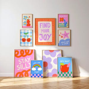 May include: A gallery wall of eight framed art prints featuring colorful illustrations of flowers, cherries, a rainbow, an apple, and stars. The prints include text such as FIND YOUR JOY, STAY SILLY, you are magic, let's read, and start with a smile.