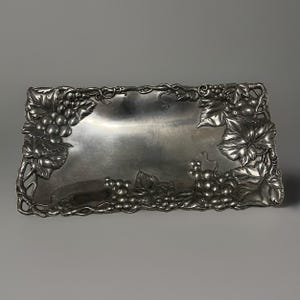May include: A rectangular silver-toned metal tray featuring a raised, embossed border design of grape clusters, vine leaves, and winding tendrils. The center of the tray is smooth and reflective, while the edges are detailed with intricate botanical motifs.