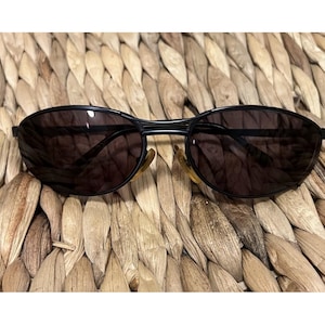 May include: Black sunglasses with dark lenses. The sunglasses have a thin, black frame and a classic oval shape. The earpieces are a light brown color. The sunglasses are resting on a woven, tan surface.