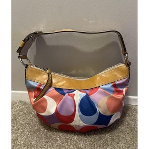 May include: A hobo-style handbag featuring a multicolored pattern of overlapping circular logos in shades of red, blue, purple, and white. The bag has a tan patent leather trim along the top edge and a matching tan leather shoulder strap.