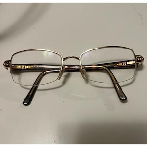 May include: Gold-tone eyeglasses with a rectangular frame and clear lenses. The glasses have a half-rim design and dark brown temples. The bridge and frame are a shiny gold color. The glasses are on a white surface.
