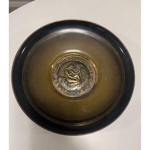 May include: A round, bronze-toned bowl with a dark rim. The center features a circular medallion with a relief of two figures and Hebrew text. The bowl has a metallic sheen and appears to be made of metal.