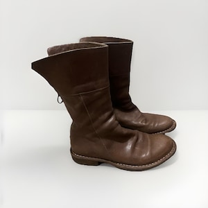 May include: A pair of brown leather boots with a worn appearance. The boots have a mid-calf height, a rounded toe, and a low heel. The leather is creased and has a distressed look, with visible stitching around the soles.