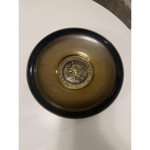 May include: A round, bronze-toned dish with a black rim. The center features a circular medallion with a figure and Hebrew text. The dish has a simple, elegant design, suitable for decorative use.