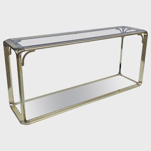 Op de afbeelding: Een rechthoekige console met een spiegelplank en een heldere glazen bovenkant. Het frame is gemaakt van gepolijst messing. De tafel heeft afgeronde hoeken en decoratieve accenten op de bovenhoeken. De tafel is ongeveer 152 cm lang.