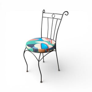 May include: A black metal chair with a round seat featuring a colorful abstract pattern in shades of blue, green, orange, and brown. The chair has a curved back and legs, and a decorative circle at the top.