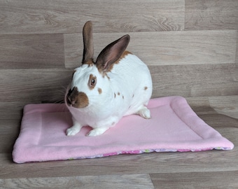 May include: A white and brown rabbit sits on a pink rectangular pet bed. The bed has a floral pattern border. The rabbit has brown ears and markings. The background is a wooden floor.