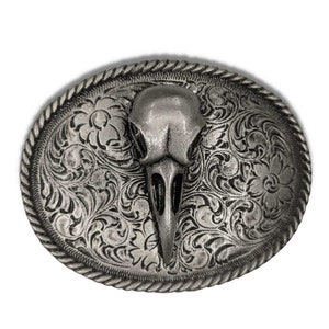 May include: A silver belt buckle with an ornate design featuring a bird skull in the center.