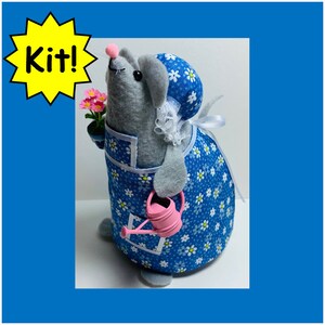 May include: A gray felt mouse wearing a blue and white floral dress and a white bonnet. The mouse is holding a pink watering can and a small pot of pink flowers.