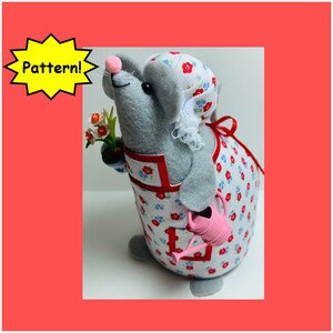 May include: A gray felt mouse wearing a white and red floral dress and a white lace bonnet. The mouse is holding a pink watering can and a small bouquet of white and yellow flowers.