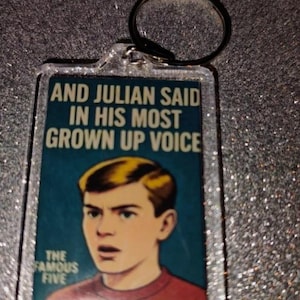 May include: A rectangular keyring with a silver ring. The keyring displays a book cover image of a young man with blond hair and the text "AND JULIAN SAID IN HIS MOST GROWN UP VOICE" and "THE FAMOUS FIVE".