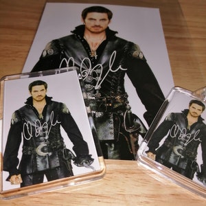 May include: An autographed photograph and two keyrings featuring a man in a black costume with a silver buckle and a necklace. The photo and keyrings have a signature in white ink.