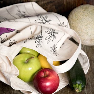 May include: A white canvas tote bag with a black floral print, filled with a red apple, two green apples, a yellow lemon, and a green zucchini.