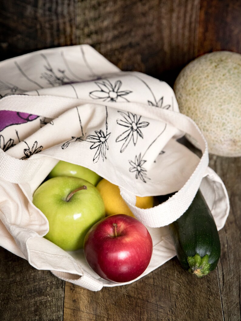 May include: A white canvas tote bag with a black floral print, filled with a red apple, two green apples, a yellow lemon, and a green zucchini.