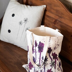May include: A white canvas tote bag with purple and black floral print and white paint splatters. The bag has two long canvas straps.