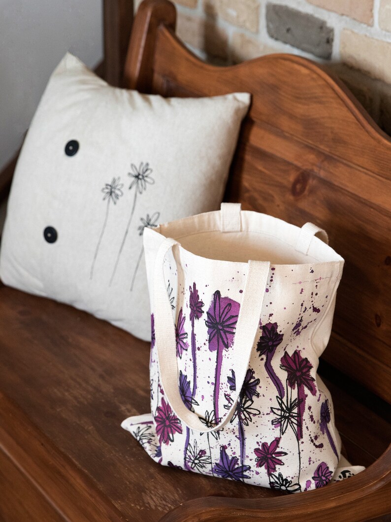 May include: A white canvas tote bag with purple and black floral print and white paint splatters. The bag has two long canvas straps.