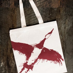 May include: A white canvas tote bag with a red ink illustration of a bird in flight. The bird is stylized with a splatter effect.