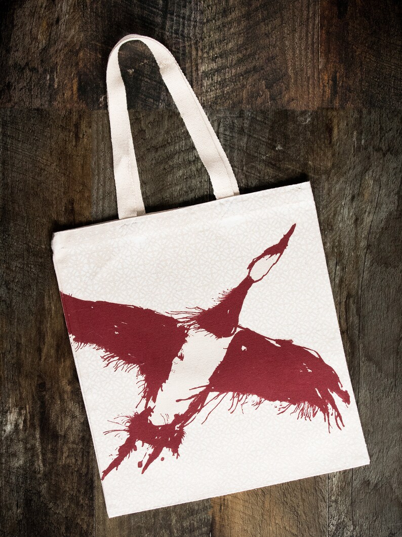 May include: A white canvas tote bag with a red ink illustration of a bird in flight. The bird is stylized with a splatter effect.