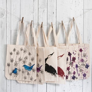 May include: Six canvas tote bags with different designs, hanging on a white wooden wall. The designs include birds, flowers, and abstract patterns in black, blue, purple, and red.