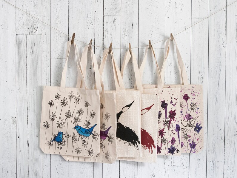May include: Six canvas tote bags with different designs, hanging on a white wooden wall. The designs include birds, flowers, and abstract patterns in black, blue, purple, and red.